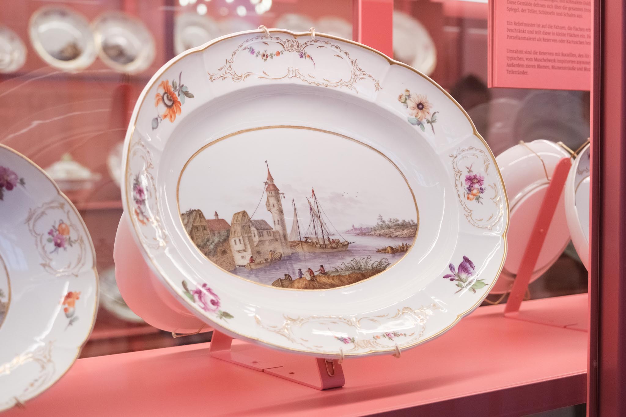 Ovale Porzellanschale mit Blumendekor und Goldrand, bemalt mit einer Hafenszene mit Turm, Booten und Figuren, in einer Museumsvitrine.