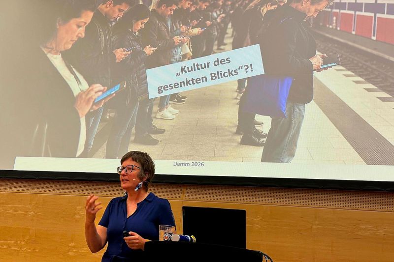 Referentin hält Vortrag vor Leinwand mit Foto wartender Menschen, die auf Smartphones blicken, und dem Titel „Kultur des gesenkten Blicks“?!