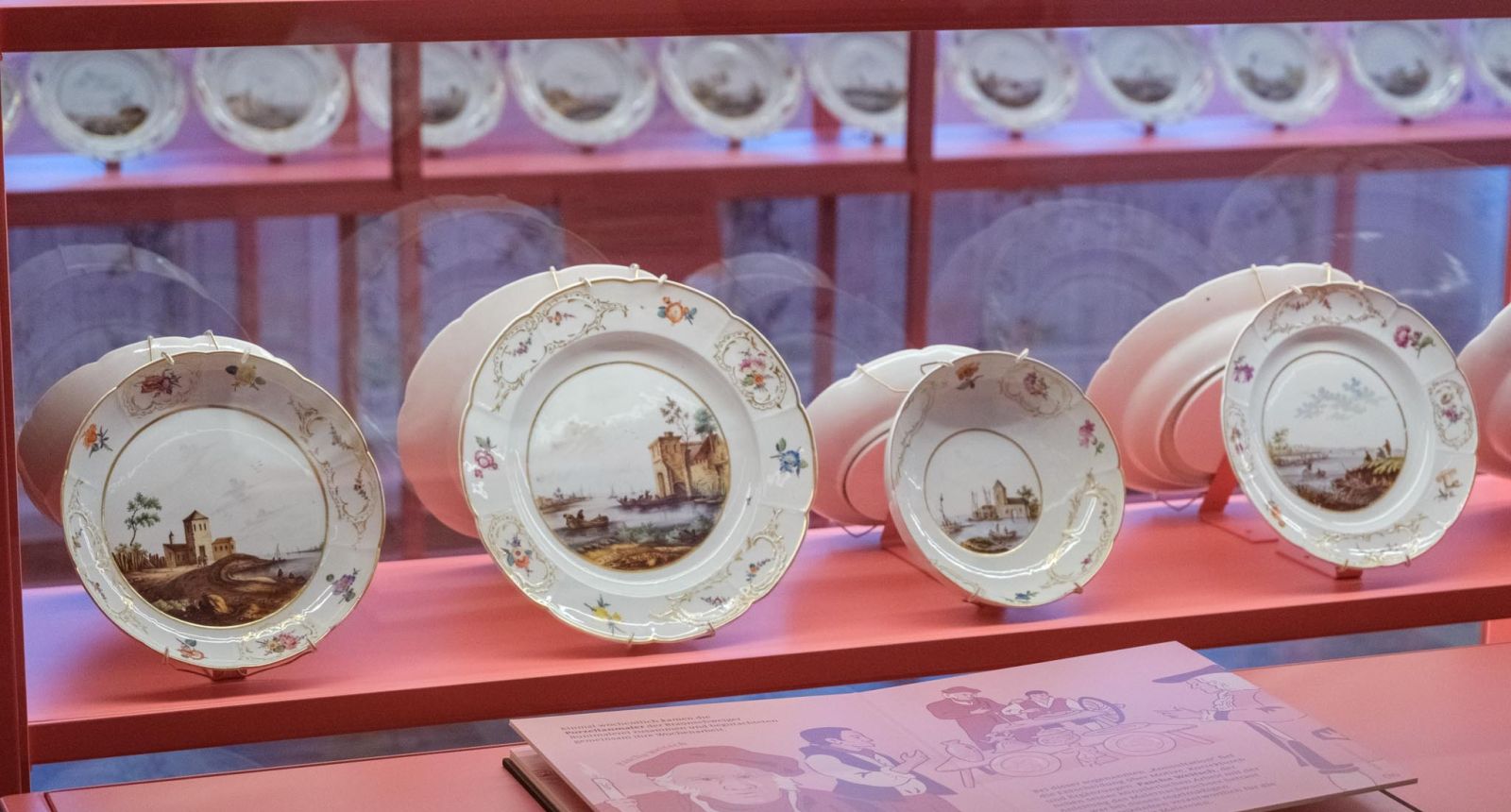 Barocke Porzellanteller mit Landschaftsmalerei und floralen Bordüren in rosafarbener Vitrine des Porzellankabinetts ausgestellt.